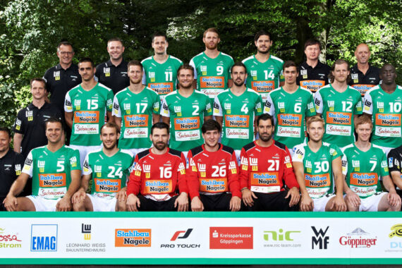 Eine Männer-Handballmannschaft stellt sich im Freien in drei Reihen auf. Sie tragen grün-weiße Trikots, an den Seiten stehen die Trainer. Zwei Spieler in der vorderen Reihe tragen rote Trikots. Am unteren Rand des Bildes sind Sponsorenlogos zu sehen.