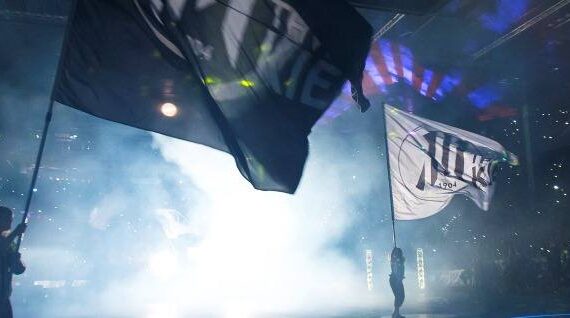 Menschen schwenken große Fahnen auf dem rauchigen, hell erleuchteten Boden einer Arena, umgeben von einer Menschenmenge und erhellt von bunten Lichtern in einer Stadionkulisse.