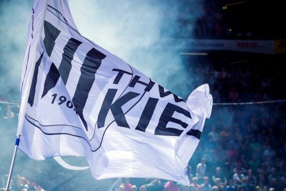 Eine große weiße Fahne mit der fettgedruckten schwarzen Aufschrift THW KIEL weht in einem mit Rauch gefüllten Stadion mit unscharfen Zuschauern im Hintergrund.