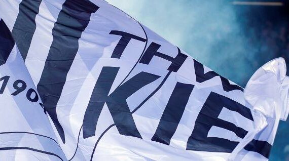 Eine große weiße Fahne mit der schwarzen Aufschrift THW KIEL 1904 wird geschwenkt, im Hintergrund sind blauer Rauch und ein überfülltes Stadion zu sehen.