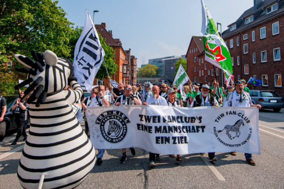 Eine Gruppe von Fans des THW Kiel marschiert mit einem großen Transparent und wehenden Fahnen eine Straße entlang, begleitet von einem Zebra-Maskottchen. Auf dem Transparent steht: "Zwei Fanclubs, eine Mannschaft, ein Ziel". Im Hintergrund sind Backsteingebäude zu sehen.