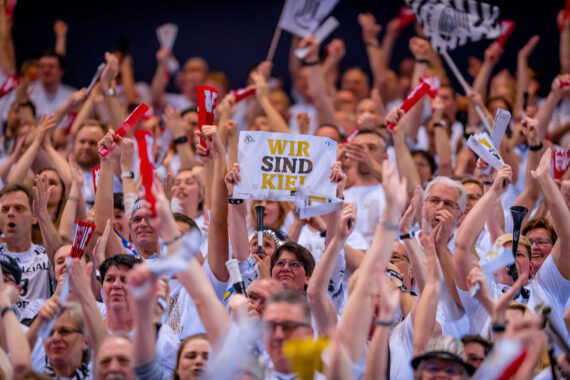 Eine große Menge begeisterter, meist weiß gekleideter Sportfans jubelt und hält Schals; eine Person in der Mitte hält ein Schild mit der Aufschrift "WIR SIND KIEL.".