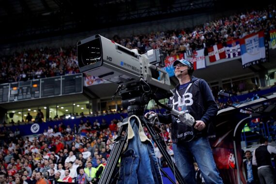 Ein Kameramann bedient eine große TV-Kamera in einer Sporthalle, in der sich viele Zuschauer versammelt haben, mit Fahnen im Hintergrund.