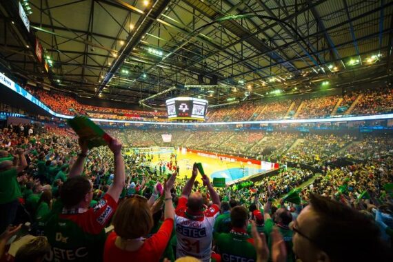 Eine große Sporthalle voller begeisterter Fans, viele in grünen Hemden, die während eines Handballspiels jubeln. Das Spielfeld und die Anzeigetafel sind hell erleuchtet, und die Tribünen sind voll mit Zuschauern.