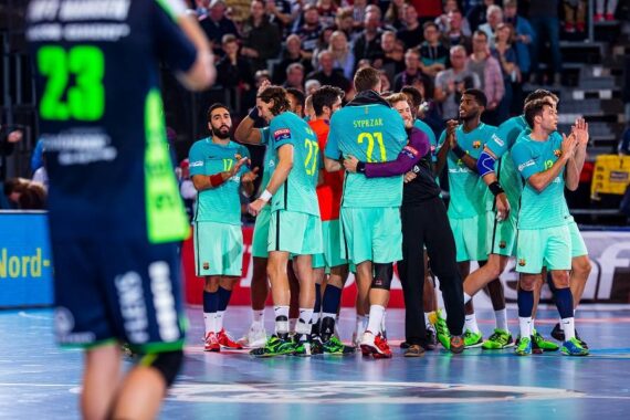 Eine Handballmannschaft in blaugrünen Trikots drängt sich auf einem Hallenplatz zusammen und jubelt. Ein weiterer Spieler in einer dunkelblauen und grünen Uniform ist unscharf im Vordergrund zu sehen. Die Zuschauer schauen von der Tribüne im Hintergrund zu.