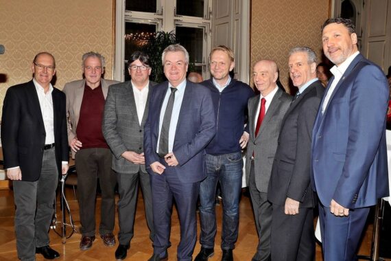 Acht Männer in formeller und halbformeller Kleidung stehen in einem warm beleuchteten, eleganten Raum mit gemusterten Tapeten und Holzböden zusammen, lächeln und posieren für ein Gruppenfoto.
