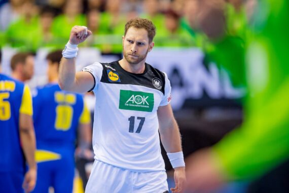Ein männlicher Handballspieler in einem weiß-schwarzen Trikot mit der Nummer 17 hebt entschlossen die Faust auf dem Spielfeld, während die Spieler in blauen und gelben Trikots im Hintergrund verschwimmen.