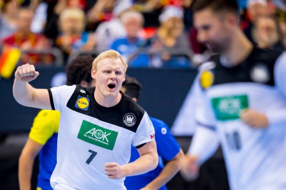Ein männlicher Handballspieler in einem weiß-schwarzen Trikot mit der Nummer 7 jubelt mit geballter Faust auf dem Spielfeld. Im Hintergrund sind andere Spieler und eine verschwommene Menge zu sehen.