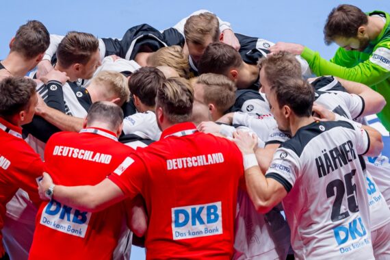 Eine Gruppe männlicher Handballspieler in rot-weißen Deutschland-Trikots steht dicht gedrängt auf einem Hallenplatz, in der Nähe steht ein Torwart in Grün.