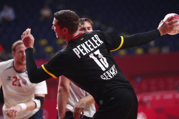 Ein Handballspieler in einem schwarzen Trikot mit Pekeler 13 Deutschland bereitet sich darauf vor, den Ball zu werfen, während er während eines Spiels von den Gegnern verteidigt wird. Der Hintergrund zeigt leere Plätze und einige unscharfe Spieler.
