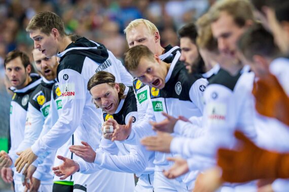 Eine Gruppe männlicher Athleten in weißen und schwarzen Trainingsanzügen feuert ihre Mannschaftskameraden bei einem Sportereignis enthusiastisch an und zeigt Begeisterung und Energie am Spielfeldrand.