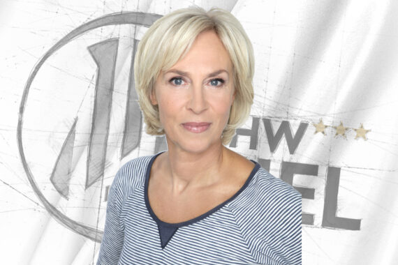 Eine Frau mit kurzen blonden Haaren, die ein blau-weiß gestreiftes Oberteil trägt, steht vor einem weißen Hintergrund, hinter dem ein graues, kreisförmiges Logo und Text teilweise sichtbar sind.