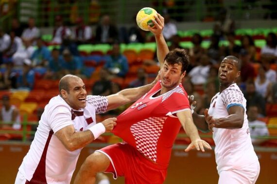Zwei Handballspieler in Aktion; der eine in einem rot-weißen Trikot wird von einem Gegner in einem weißen Trikot am Trikot gezogen, während ein anderer Spieler in Weiß zusieht. Die Szene ist dynamisch und intensiv, in einer Halle.