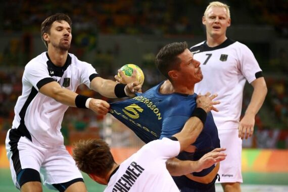 Ein Handballspieler in einem blauen Trikot wird während eines Spiels von einem Gegner in einem weißen Trikot gewaltsam am Hemd gepackt; ein anderer Spieler in Weiß fällt, und ein dritter steht in der Nähe und schaut zu.