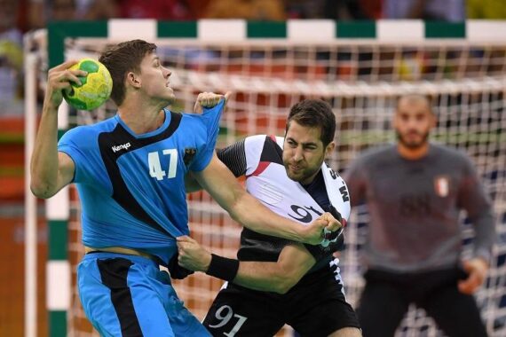 Zwei männliche Handballspieler liefern sich einen intensiven Wettkampf; der eine im blauen Trikot (#47) wird während eines Spiels von einem Gegner im schwarz-weißen Trikot (#91) am Trikot gezogen; im Hintergrund ist ein Torwart zu sehen.