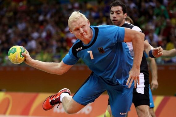 Ein männlicher Handballspieler in einem blauen Trikot bereitet sich darauf vor, den Ball zu werfen, während er von einem Gegner in einem schwarz-weißen Trikot während eines Spiels eng verteidigt wird. Das Publikum ist im Hintergrund verschwommen.