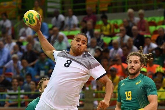 Ein männlicher Handballspieler in einem weiß-grauen Trikot springt, um den Ball zu werfen, während ein Spieler in einem grünen Trikot zusieht. Im Hintergrund sind unscharfe Zuschauer in einem Stadion zu sehen.