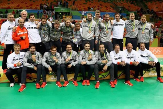 Die deutsche Volleyballmannschaft der Männer posiert mit ihren Bronzemedaillen bei den Olympischen Spielen 2016 in Rio, lächelnd und mit ihren Medaillen in der Hand, gekleidet in passende grau-weiße Deutschland-Jacken.