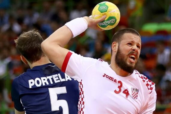 Ein männlicher Handballspieler im weiß-roten Kroatien-Trikot bereitet sich auf den Wurf vor, während ein Spieler in einem blauen Trikot mit der Portela 5 auf dem Rücken den Ball abwehrt. Die Aktion findet in einer Halle statt.