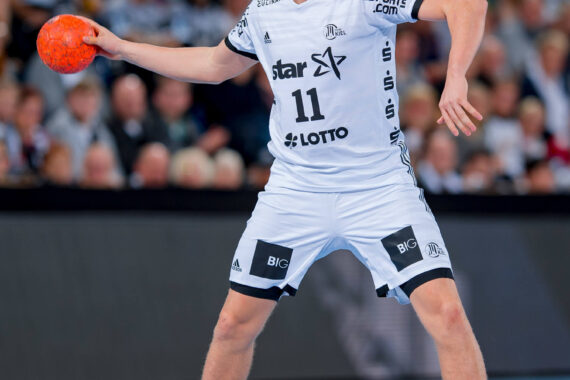 Ein männlicher Handballspieler, der ein weißes Trikot mit der Nummer 11 trägt, bereitet sich darauf vor, während eines Spiels einen orangefarbenen Ball zu werfen, wobei das Publikum im Hintergrund unscharf zu sehen ist.