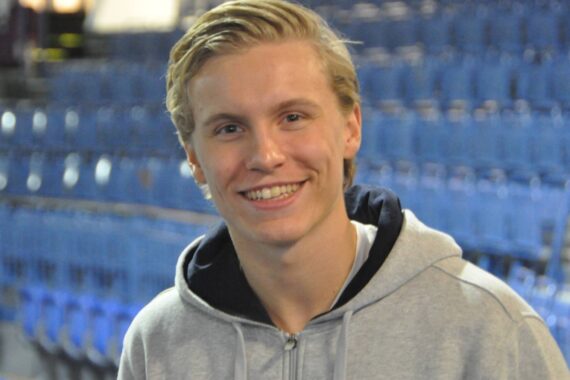 Ein junger Mann mit blonden Haaren und einem grauen Kapuzenpulli lächelt in die Kamera. Er befindet sich in einem Innenraum, und im Hintergrund sind blaue stadionähnliche Sitze zu sehen.