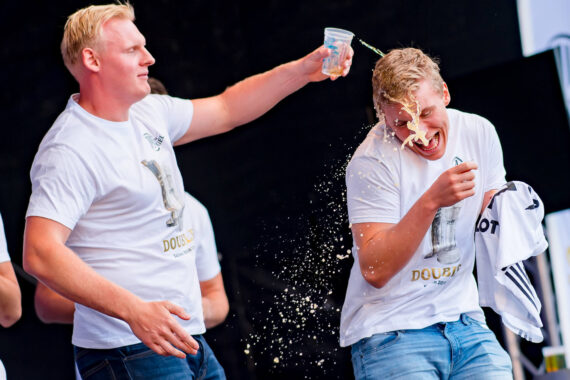 Ein Mann schüttet spielerisch ein Getränk über den Kopf eines anderen Mannes, so dass dieser lacht und sein eigenes Getränk verschüttet. Beide tragen weiße T-Shirts und Jeans, und die Szene ist lebhaft und fröhlich.