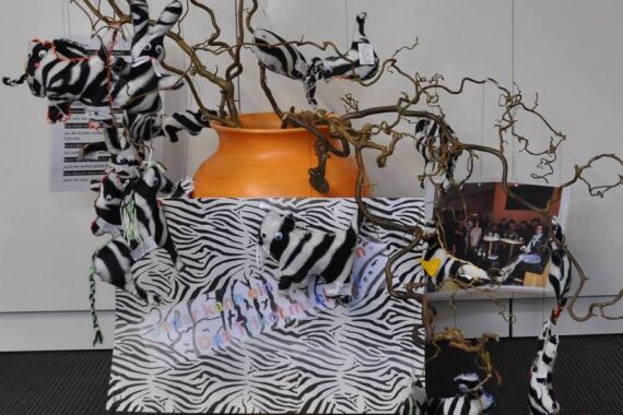 In einer orangefarbenen Vase befinden sich verschlungene Zweige, die mit kleinen Plüschzebras verziert sind, ein Poster mit Zebramuster und ein Gruppenfoto. Die Auslage steht vor einer weißen Wand auf einem dunklen Boden.