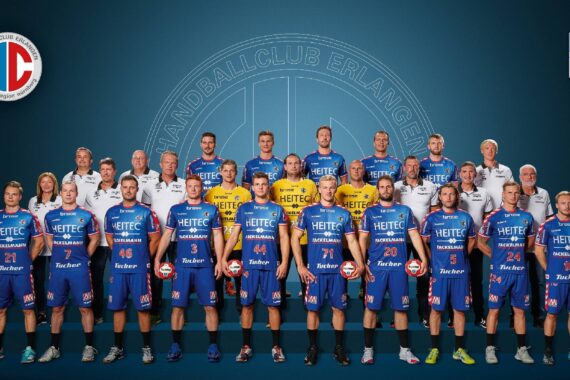 Eine Herren-Handballmannschaft und ihre Betreuer posieren gemeinsam in blauen Trikots vor einem blauen Hintergrund mit dem Logo des Handballclubs Erlangen und Sponsorenlogos. Die Spieler halten Handbälle und stehen in zwei Reihen.