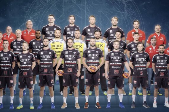 Eine Männerhandballmannschaft in schwarzen Trikots steht in zwei Reihen und hält Handbälle in der Hand, auf beiden Seiten stehen Trainer in roten Hemden, alle schauen nach vorne vor einem blauen Hintergrund mit einem schwachen kreisförmigen Logo.