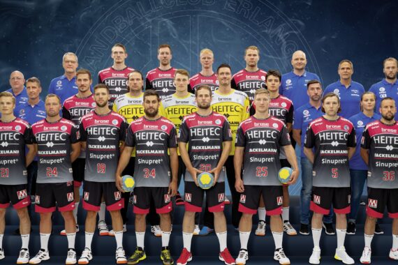 Eine Handballmannschaft der Männer posiert für ein Gruppenfoto in Uniform und mit Handbällen in der Hand. Die Spieler stehen vorne, Trainer und Betreuer stehen hinter ihnen, alle schauen nach vorne vor einem dunkelblauen Hintergrund mit einem schwachen Logo.