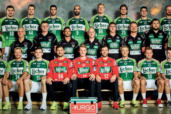 Eine Herren-Handballmannschaft stellt sich in grün-weißen Trikots für ein Gruppenfoto auf, die Trainer stehen hinter ihnen. Vorne sitzen drei Torhüter in roten Trikots, in der Mitte steht ein Verbandskasten mit der Aufschrift URGO.