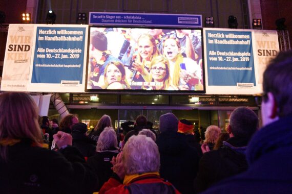 Eine Menschenmenge schaut auf eine große Leinwand im Freien, die jubelnde Fans bei einem Handball-Event zeigt. Banner über der Leinwand heißen die Besucher des Handballbahnhofs zu den deutschen Spielen vom 10. bis 27. Januar 2019 willkommen.