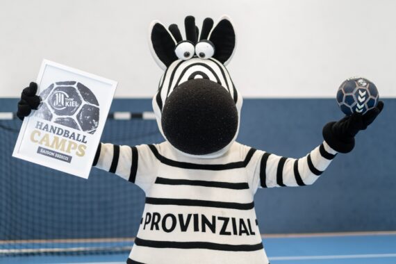 Eine Person in einem Zebra-Maskottchenkostüm hält einen Handball und ein Schild mit der Aufschrift Handball Camps hoch. Im Hintergrund ist ein blauer Hallensportplatz mit einem Torpfosten zu sehen. Das Maskottchen trägt die Aufschrift PROVINZIAL auf seinem Trikot.