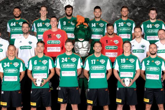 Eine Männer-Handballmannschaft posiert in einer Halle in grün-weißen Trikots, dahinter stehen Trainer und Betreuer in weißen Hemden. Die Spieler halten Handbälle in der Hand, und ein grauer felsiger Hintergrund ist zu sehen.
