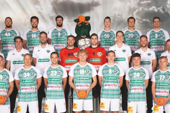 Eine Männer-Handballmannschaft posiert für ein Gruppenfoto in grün-weißen Trikots mit Trainern und Mitarbeitern; zwei Spieler halten orangefarbene Handbälle, und ein grünes Maskottchen steht in der hinteren Reihe. Stürmischer Stadionhintergrund hinter ihnen.