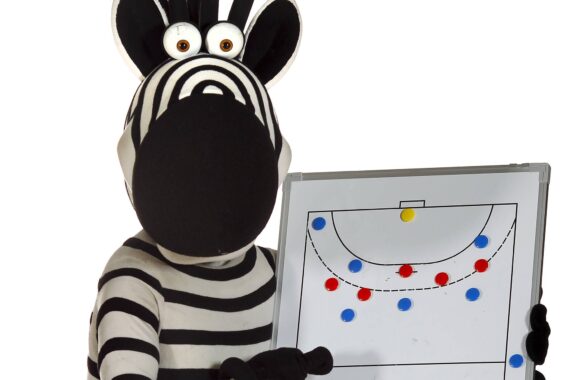 Eine Person im Zebrakostüm hält ein Whiteboard, auf dem ein Diagramm einer Fußballstrategie mit roten und blauen Markierungen auf einem Torraum abgebildet ist.