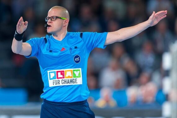 Ein Schiedsrichter in einer blauen Uniform mit der Aufschrift "Play for Change" auf der Vorderseite signalisiert während einer Sportveranstaltung mit beiden Armen in entgegengesetzte Richtungen.