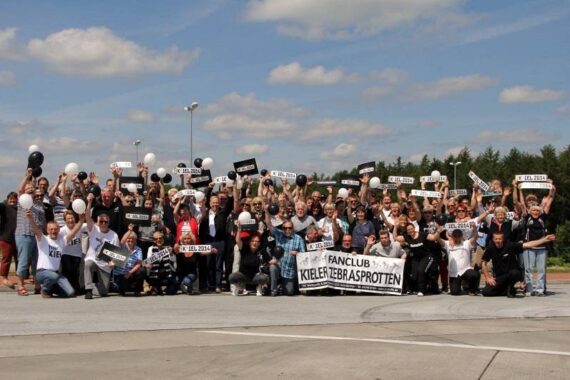 Eine große Gruppe von Menschen im Freien hält Schilder, Luftballons und ein Banner mit der Aufschrift Fanclub Kieler Sprotten an einem sonnigen Tag, mit Bäumen und Wolken im Hintergrund.