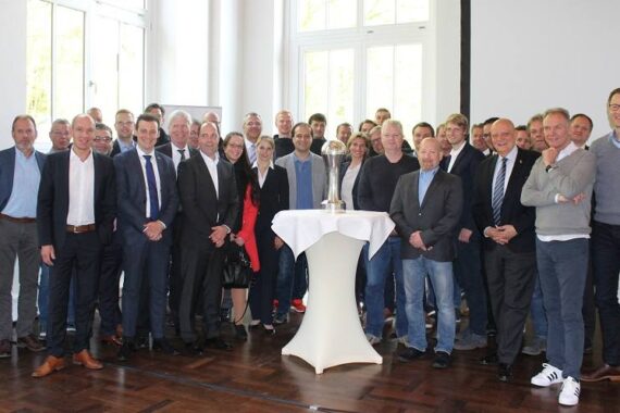 Eine Gruppe von etwa 30 Personen, meist Männer in Business- oder eleganter Freizeitkleidung, steht in einem hellen Raum mit großen Fenstern um einen hohen runden Tisch herum und lächelt in die Kamera.