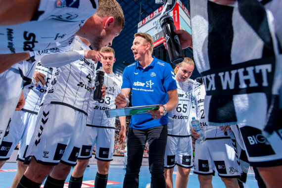Ein Handballtrainer in einem blauen Hemd gibt seiner Mannschaft, die weiße Trikots trägt, während einer Auszeit Anweisungen. Die Spieler hören mit Wasserflaschen in der Hand auf einem Hallenplatz aufmerksam zu.