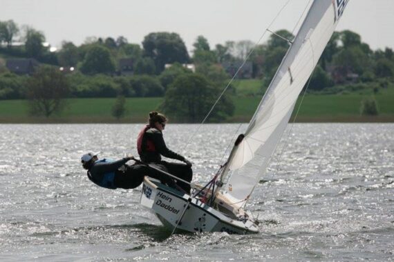 Zwei Personen fahren mit einem kleinen Boot über einen See und lehnen sich über den Rand, um das Gleichgewicht zu halten, während das Boot kippt. Das Wasser ist leicht kabbelig, und im Hintergrund ist ein grünes, von Bäumen gesäumtes Ufer zu sehen.