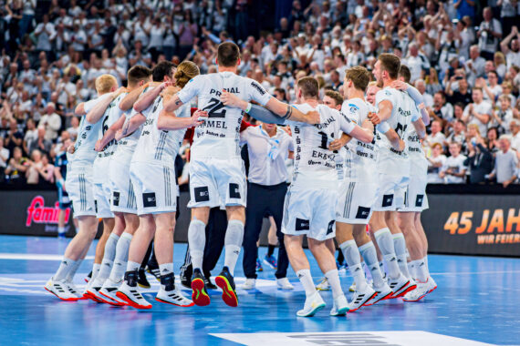 Eine Herren-Handballmannschaft in weißen Trikots feiert auf einem blauen Hallenplatz, die Arme umeinander gelegt, umgeben von jubelnden Fans auf der Tribüne.