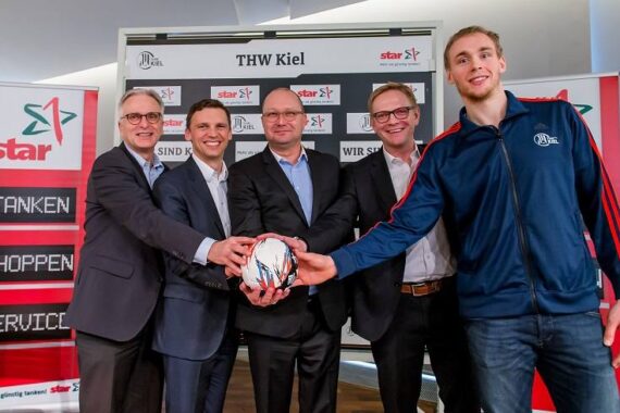 Fünf Männer in Business- und Sportkleidung stehen lächelnd auf einer Presseveranstaltung und halten einen silberglänzenden Ball in der Hand. Im Hintergrund sind die Logos des THW Kiel und der Stars auf Bannern zu sehen.