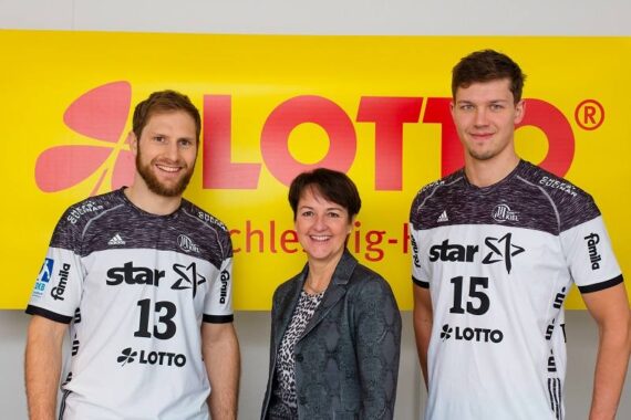 Zwei männliche Sportler in weißen Trikots mit den Nummern 13 und 15 stehen zu beiden Seiten einer lächelnden Frau in Geschäftskleidung vor einem gelben LOTTO-Banner.