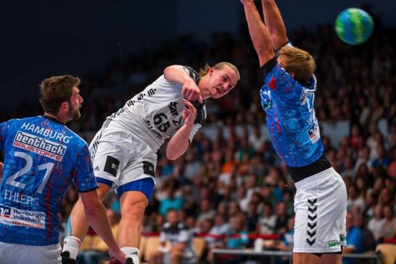 Ein Handballspieler in einem weißen Trikot springt, um den Ball an zwei Verteidigern in blauen Trikots vorbei zu schießen, während im Hintergrund eine unscharfe Menschenmenge zu sehen ist.
