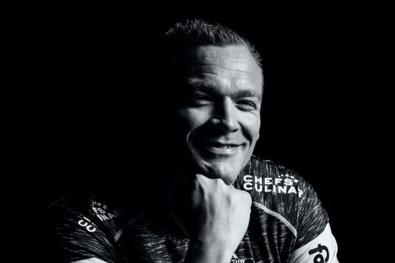 Ein Mann mit kurzen Haaren, der ein Sporttrikot trägt, lächelt und stützt sein Kinn auf seine Hand. Das Bild ist in Schwarz-Weiß mit dramatischer Beleuchtung, die tiefe Schatten auf sein Gesicht wirft.