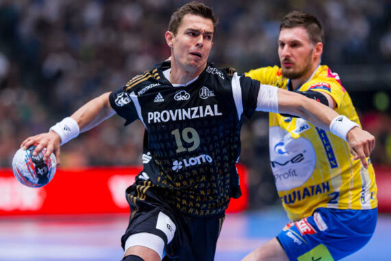 Zwei männliche Handballspieler in Aktion auf einem Spielfeld; der Spieler in Schwarz versucht zu schießen oder zu passen, während er von einem Spieler in Gelb eng verteidigt wird. Im Hintergrund ist ein unscharfes Publikum zu sehen.