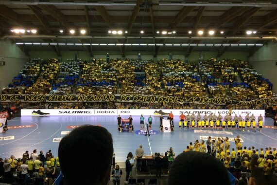Eine überfüllte Sporthalle mit Fans in Gelb und Blau, ein großes polnisches Banner auf der Tribüne und zwei Mannschaften, die sich vor einem Handballspiel auf dem blauen Spielfeld aufstellen.