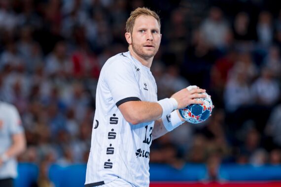 Ein männlicher Handballspieler in einer weißen Uniform hält einen bunten Handball und schaut konzentriert. Er steht auf einem Hallenplatz mit einer unscharfen Menschenmenge im Hintergrund.