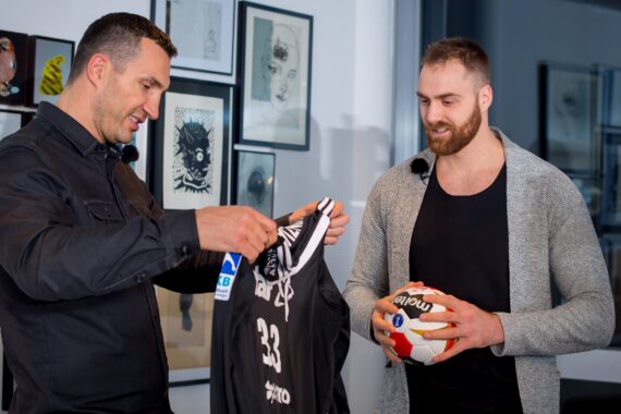 Zwei Männer stehen in einem Haus; der eine hält ein schwarzes Sporttrikot in der Hand, während der andere einen Handball in der Hand hält und lächelt. Hinter ihnen hängen gerahmte Kunstwerke an der Wand.
